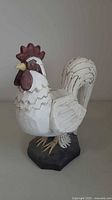 Side angle view of the white painted wooden rooster figurine with red comb and wattle, showing carved feather details and attached to an octagonal black base.