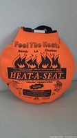 Photo shows bright orange HEAT-A-SEAT heated seat cushion with black printed instructions and branding. Carrying handle visible on top.