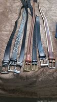 Seven men's XL belts laid out on a brown surface showing different styles and materials including leather, braided, and fabric with various buckles.