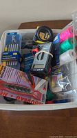 Plastic container with assorted tools including a measuring tape, utility knife, spools of colored thread or wire, screwdriver, small hand tools, and a digital multimeter