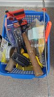 Photo showing mixed lot of 11-15 assorted hand tools in a blue basket including hammer, saw, voltage tester, sanding block, wire spool, and other tools.