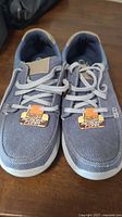 Pair of men's Skechers blue-gray mesh lace-up shoes on wooden surface, showing front and top views with tags attached about Memory Foam cushioning.