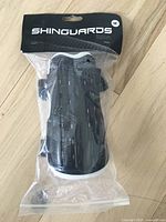 Black soccer shin guards in original sealed packaging labeled 'SHINGUARDS Sport' with medium size indicator.