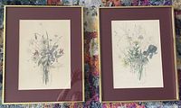 Pair of framed floral lithographs with burgundy mats and brass frames, resting on multicolored carpet.