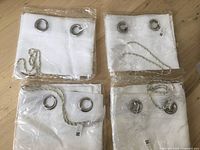Four white sheer curtains in original plastic packaging, each folded showing metal grommets and decorative cords on wooden floor.