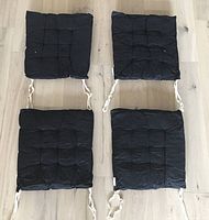 Four black seat cushions laid out flat on wooden floor showing tufted grid pattern and cream fabric ties.