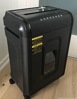 Front and angled view of the black AmazonBasics shredder showing the micro-cut slot, safety and function icons, and the pullout basket window on the front.