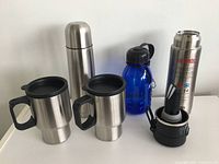 Set of one stainless steel thermos, two travel mugs, and one blue water bottle arranged on a white surface.