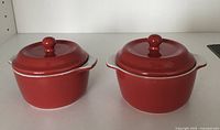 Two red ceramic mini casseroles with lids, side by side on white surface.
