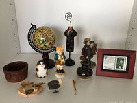 Overview of vintage figurines and decorative items arranged on a shelf, including globe, wooden bowl, figurines, framed card, and small collectibles.
