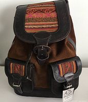 Front view of backpack showing dark brown suede base, black leather trim and buckles, and colorful woven fabric on front flap and pockets.