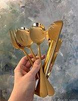Hand holding all four gold-tone metal serving utensils together, showing the fork, slotted spoon, solid spoon, and tongs with polished gold finish.