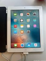 Front view of Apple iPad with screen on, showing home screen icons, white bezel, charging cable connected at bottom.