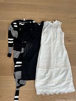 Three women's dresses laid flat on a wooden surface: a white sleeveless lace dress, a solid black dress, and a black dress with white striped pattern and tie belt at the waist.