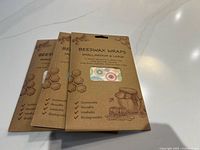 Three unopened packs of beeswax wraps arranged side by side on a white counter, showing front packaging with product text and a window revealing colorful wrap patterns.