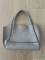 Front view of taupe pebbled leather handbag with double carry handles and black piping detail on wood floor