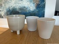 Three off-white/cream ceramic planters displayed on wooden surface; one with three legs, two cylindrical.