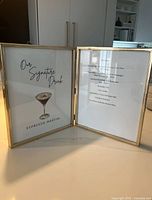 Photo of double brass picture frames standing open on a light surface with kitchen background. Holds papers with text and a martini image.
