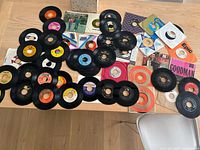 Top view of about 46 vintage 45 RPM vinyl records spread out on a wooden table, showing various labels and some paper sleeves.