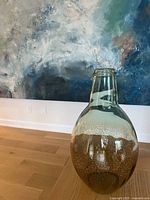 Front view of tall clear glass vase with speckled amber design covering the bottom half; vase placed on wooden floor with blue abstract painting in background.