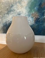 Front view of the large white glass vase with a glossy surface placed on a wooden surface and abstract blue wall art in the background.
