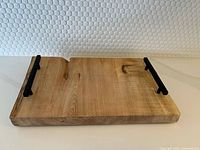 Front view of live edge maple wood serving tray with black metal handles placed on a white surface against textured white wall.