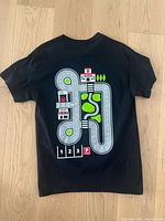 Back view of new men's black cotton t-shirt with colorful road graphic including gas station, workshop, parking spots, trees, and a pond.
