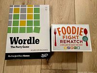 Two party board games boxes: Wordle The Party Game (white box with colored grid) and Foodie Fight Rematch (silver box with orange lettering and fork and spoon graphics).