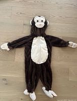 Front view of a children's monkey costume laid flat on wooden floor showing attached hood with monkey face, zip front zipper, plush brown faux fur material with white belly panel, mittens and foot covers shaped like monkey hands and feet.