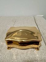 Closed rectangular porcelain trinket box with shiny gold-tone finish and shaped lid with gentle curves, sitting on a cushioned surface.