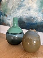 Front view of both vases side by side, showing green glass vase with round bottom and narrow neck, and rustic speckled earthenware vase.