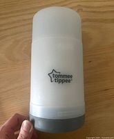 White and grey cylindrical travel bottle warmer with Tommee Tippee logo shown on wooden surface held in hand