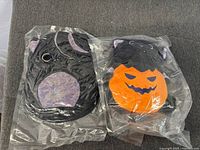 Two factory-sealed Halloween plush toys, cat and pumpkin designs, on grey surface