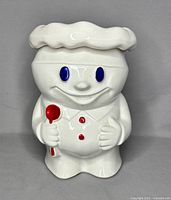 Front view of white ceramic Pillsbury Doughboy cookie jar showing blue eyes, red spoon and buttons