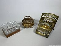 Three items displayed side by side: a glass trinket dish on brass base, small brass trinket dish with handle and floral motifs, and foldable brass letter rack with floral pattern and stamp compartment.