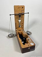 Antique balance scale with wooden box, showing balance arms and gold-tone pans hanging by chains, one chain broken, with compartments for weights and some weights inside.
