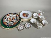 Full view of assorted china pieces including Aynsley sugar bowl and creamer, Schumann fruit motif plates, Queen Anne creamer and sugar bowl, several small cups and plates with floral patterns and gold accents.
