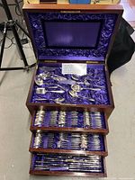 Open wooden chest showing top compartment and three drawers with purple satin lining and assorted flatware.