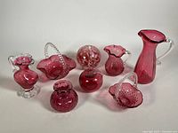 Full view of eight cranberry glass pieces arranged in two rows showing pitchers, trinket dishes, orb, and apple decor piece