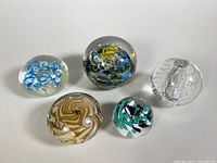 Five glass paperweights arranged on a white surface showing variety of colors and embedded designs