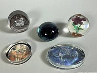 Five glass paperweights arranged on a surface, including two large oval art reproduction paperweights and three spherical orbs (some with chips near base).