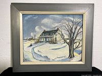 Framed canvas painting showing a snow-covered rural house and bare tree in the foreground under cloudy sky. Close view of frame corners.