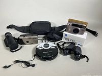 Minolta RIVA Zoom 70 Date camera in silver with case and original box, Bausch and Lomb 8x24 binoculars, and Studebaker compact portable CD player with headphones and case arranged together on a light surface.
