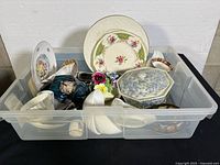 Plastic bin filled with assorted porcelain and china pieces including plates and decorative items. Visible delicate floral designs and some figurines.