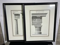 Two framed Roman architectural prints leaning against a white wall. One print shows a Roman architectural column detail, the other print shows side-by-side Grecian and Roman Doric columns. Both have black wood frames, mats, and glass fronts.