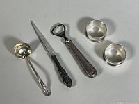 Full view of five sterling silver items: two napkin rings, small ladle, letter opener, bottle opener laid out on white surface.