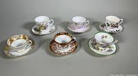 Six teacups and saucers arranged in two rows on a gray surface showing variety of floral and gold designs