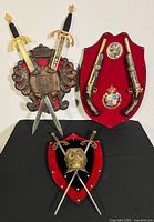 Three metal and wood coats of arms displayed on a black surface, showing details of swords, pistols, and chest plate.