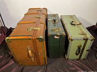 Three vintage steamer trunks side by side showing relative sizes and wear, placed on dark fabric