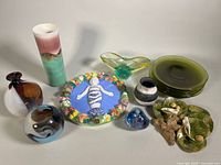 Wide shot showing assortment of seven green glass side plates stacked, various glass and ceramic vases including tall gradient vase, two small multicolored vases, and ceramic decorative plates with fruit and figurine.